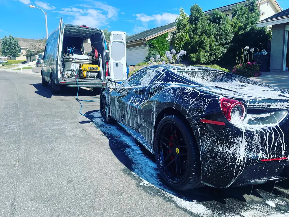The MVP van parked at the curb with its doors open, water tank and generator inside, next to a foamed-up Ferrari